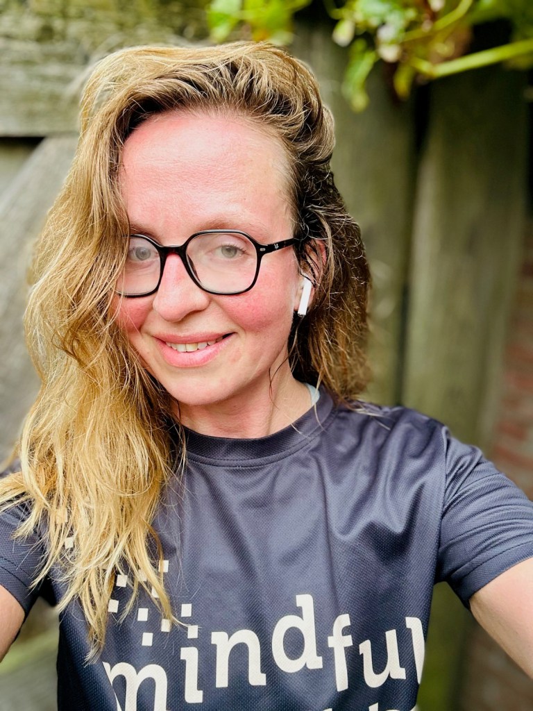 Een vrouw met lang, golvend haar en een zwart shirt met het woord 'mindful' erop, die glimlacht terwijl ze een selfie neemt in een groene omgeving.