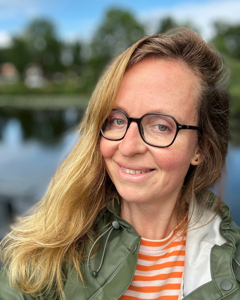 Een vrouw met lange, golvende haren en een bril, die glimlacht voor een natuurlijke achtergrond met water en bomen.