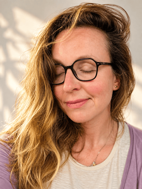 Een vrouw met een zachte glimlach en gesloten ogen, met lang golvend haar en een paar zwarte bril, gefotografeerd in natuurlijk licht.
